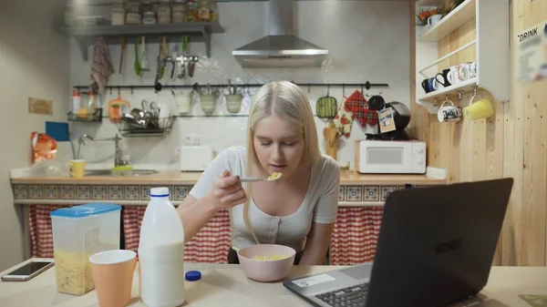 Genç bir kadın dizüstü bilgisayardan video izliyor ve mısır gevreği yiyor..