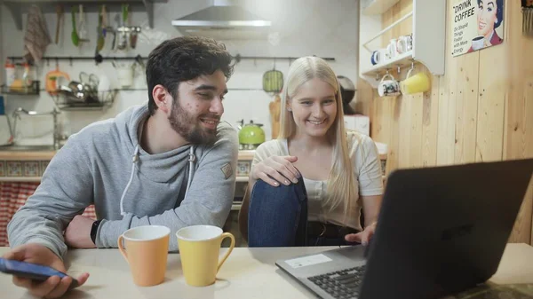 Young couple talking and using modern laptop and phone in the kitchen. — Stock Photo, Image