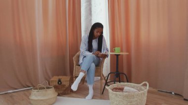 Genç bir kadın evdeki rahat bir odada otururken kahve içer ve telefon kullanır.