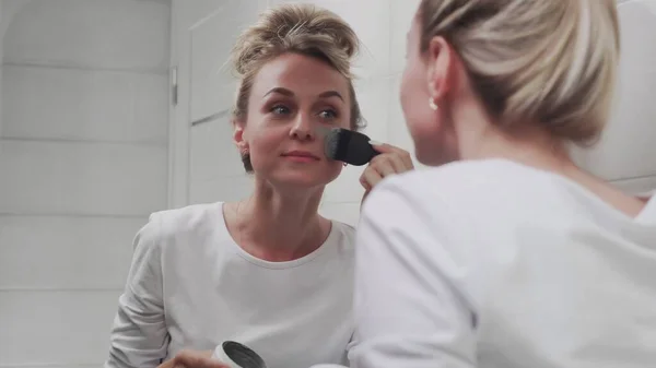 女人用刷子在脸上涂上黏土面罩,照镜子 — 图库照片
