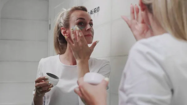 Mujer aplicando máscara de arcilla en su cara mirando en el espejo —  Fotos de Stock