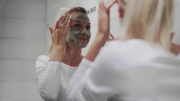 Wanita cantik mengenakan masker tanah liat di wajahnya tampak di cermin — Stok Foto