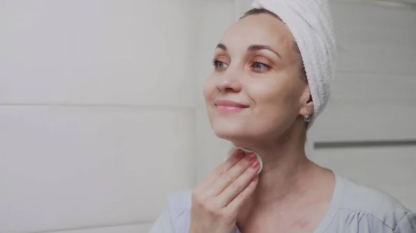 Mujer adulta con una toalla en la cabeza aplicando loción y mirando en el espejo disfrutar de la belleza natural y saludable — Foto de Stock