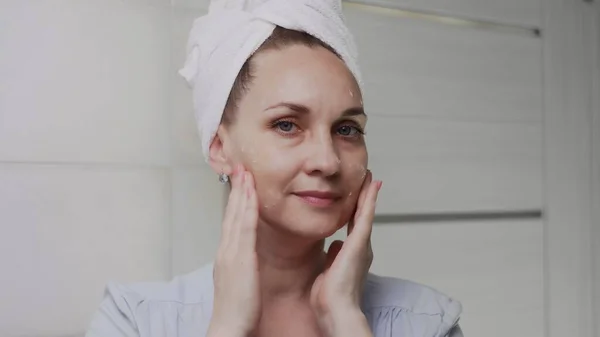 Mujer adulta con una toalla en la cabeza aplicando crema y mirando en el espejo disfrutar de la belleza natural y saludable — Foto de Stock