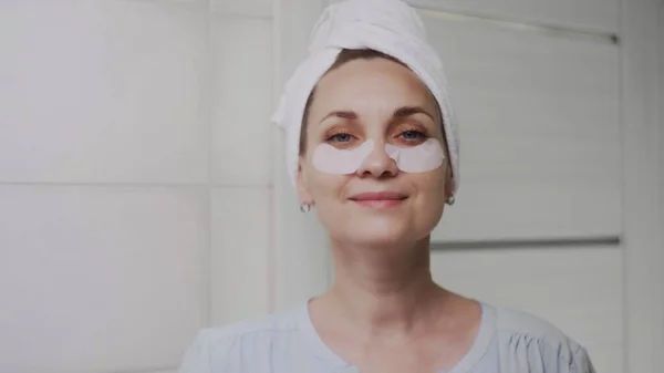 Acercamiento de la mujer adulta con una toalla en la cabeza y almohadillas de colágeno debajo de los ojos y mira a la cámara como en el espejo — Foto de Stock