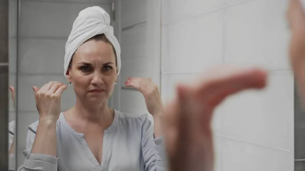 Frustrated Adult woman with a towel on her head looks at her face in the bathroom mirror — Stock Photo, Image