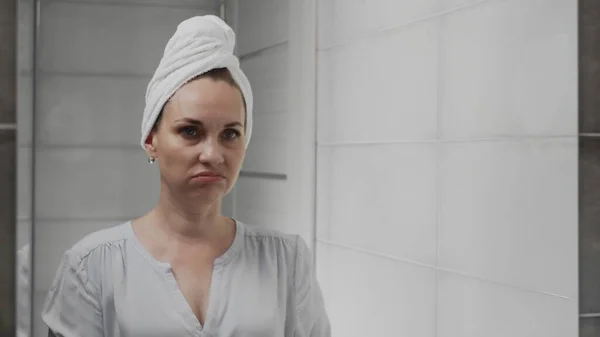Frustrated Adult woman with a towel on her head looks at her face in the bathroom mirror — Stock Photo, Image