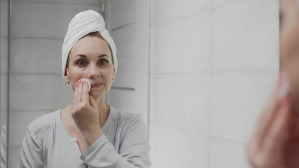 Взрослая женщина с полотенцем на голове применяя лосьон и глядя в зеркало наслаждаться природной красотой и здоровым — стоковое фото