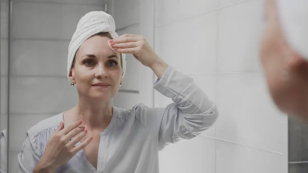 Mujer adulta con una toalla en la cabeza aplicando loción y mirando en el espejo disfrutar de la belleza natural y saludable — Foto de Stock