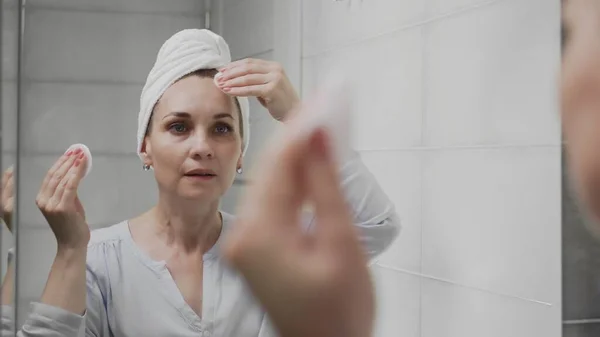 Mulher adulta com uma toalha na cabeça aplicando loção e olhando no espelho desfrutar de beleza natural e saudável — Fotografia de Stock