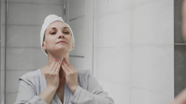 Mujer adulta con una toalla en la cabeza aplicando loción y mirando en el espejo disfrutar de la belleza natural y saludable — Foto de Stock