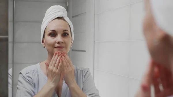 Mujer adulta con una toalla en la cabeza aplicando crema y mirando en el espejo disfrutar de la belleza natural y saludable — Foto de Stock