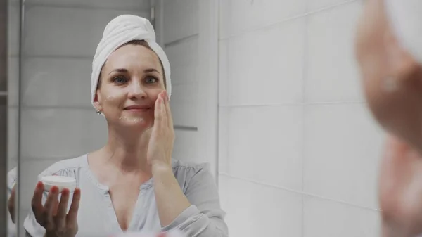 Взрослая женщина с полотенцем на голове нанесения крема и глядя в зеркало наслаждаться природной красотой и здоровым — стоковое фото