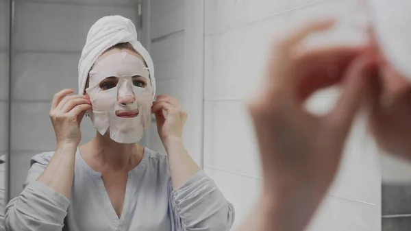 Mujer adulta con una toalla en la cabeza que se aplica una máscara en la cara y mira a la cámara — Foto de Stock