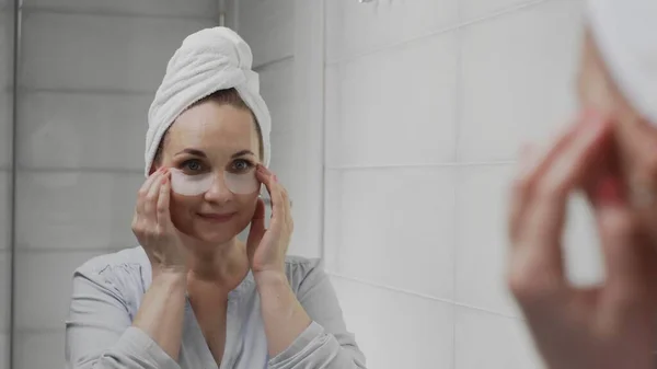 Adult woman with a towel on her head puts collagen pads under her eyes and looks in the mirror — Stock Photo, Image