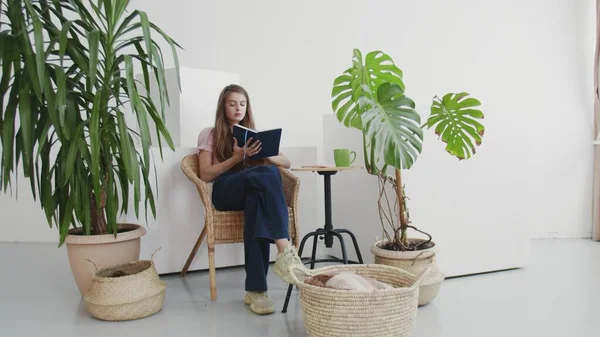 Schöne Frau liest ein Buch in ihrem Wohnzimmer zu Hause. Entspannte junge Frau sitzt neben Zimmerpflanzen — Stockfoto