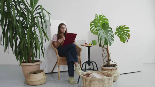 Schöne junge Frau sitzt auf Stuhl und malt ein Bild in ihrem heimischen Atelier. — Stockfoto