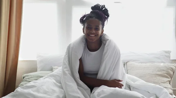 Young happy african woman sits om bed, looks at the camera and smiles — Stock Photo, Image