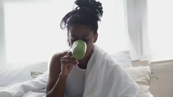 Close up of Young African American Woman sits in bed, drinks morning coffee and uses phone — Stock Photo, Image