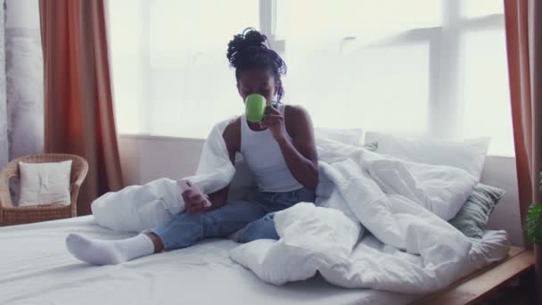 Pretty Young donna afroamericana si siede a letto, beve caffè del mattino e utilizza il telefono — Video Stock