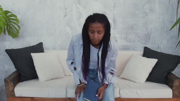 Young african american woman sits sofa and reads a book — Stock Video