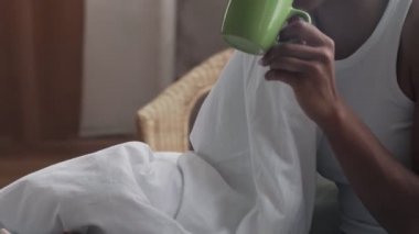 Afro-Amerikalı bir kadın yatakta oturur, sabah kahvesini içer ve telefon kullanır.