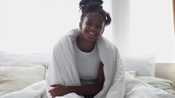 Joven mujer africana feliz se sienta om cama, mira a la cámara y sonríe — Vídeo de stock