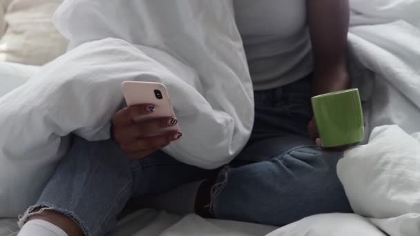 Молодая афроамериканка сидит в постели, пьет утренний кофе и пользуется телефоном — стоковое видео