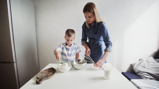 Család a konyhában. Gyönyörű anya kisfiúval. A kisfiú egy kanál lisztet tölt a fémtálba, az anyja pedig egy csésze tejet. — Stock videók