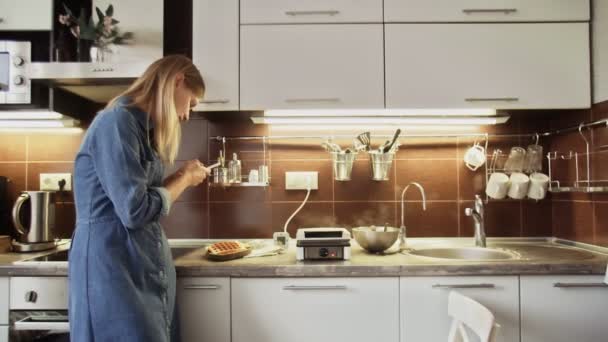 Веселая женщина фотографирует еду на смартфоне на кухне. Милая девушка делает фото вафель на столе дома. Блогер продуктов питания с помощью мобильного телефона на кухне — стоковое видео