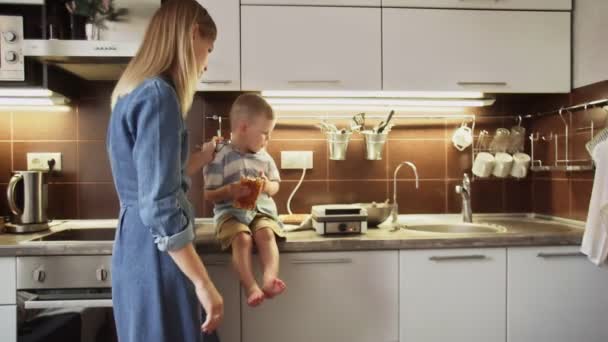 Die junge Mutter und ihr Sohn backen in der heimischen Küche Waffeln. Kleiner Junge sitzt auf dem Tisch und isst die Waffel — Stockvideo