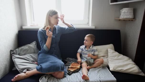 若いです母親と彼女の息子はソファに座って、ワッフルを食べ、ジュースを飲みます — ストック動画