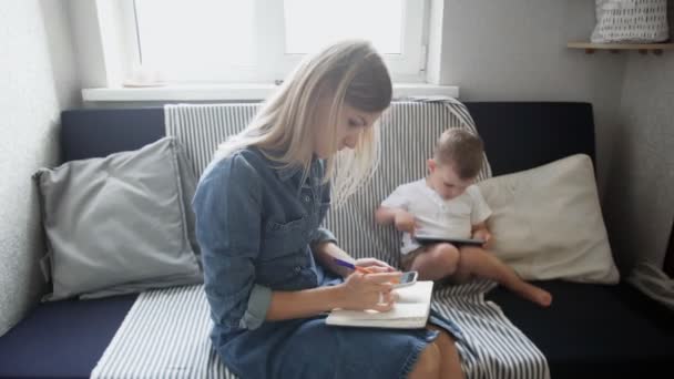 Junge Frau mit kleinem Kind, die von zu Hause aus arbeitet. Mutter arbeitet, während ihr Sohn Cartoon auf Tablet guckt — Stockvideo