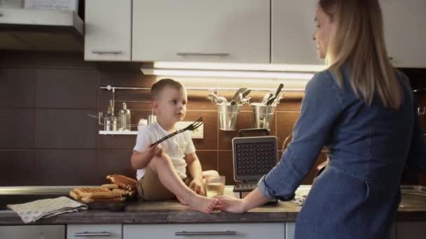 Die junge Mutter und ihr Sohn backen in der heimischen Küche Waffeln. Kleiner Junge sitzt auf Tisch und trinkt Saft — Stockvideo