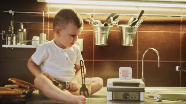 Młody chłopiec robi gofry w maszynie do gofrów w domowej kuchni. Mały chłopiec siada na stole. Wkłada zegar na gofrownicę. — Wideo stockowe