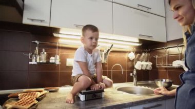 Küçük çocuk mutfakta waffle makinesinin üzerine oturur.