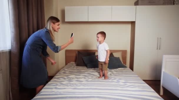 Šťastný malý chlapec skákání na rodiče postel v ložnici, zatímco jeho matka je nahrávání videa na telefonu
