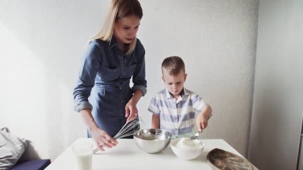Семья на кухне. Прекрасная мать с маленьким сыном. Мальчик наливает ложку муки в металлическую миску, а его мать наливает чашку молока. — стоковое видео