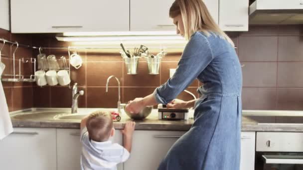 Νεαρή μητέρα φτιάχνει βάφλες σε βαφλιέρα ενώ ο γιος της παίζει με παιχνίδι στην κουζίνα του σπιτιού — Αρχείο Βίντεο