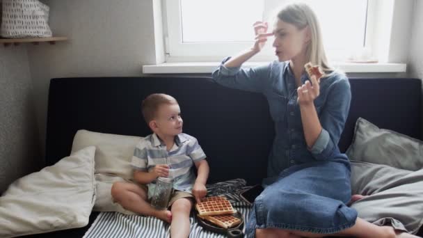 Junge Mutter und ihr Sohn sitzen auf Couch, essen Waffeln und trinken Saft — Stockvideo