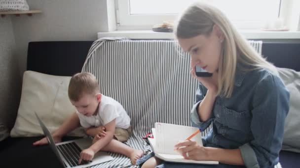 Wanita sibuk berbicara di telepon dan membuat catatan sementara anaknya bermain dengan notebook — Stok Video