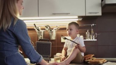 Genç anne ve oğlu ev mutfağında waffle makinesinde waffle yapıyorlar. Küçük çocuk masaya oturur ve meyve suyu içer.