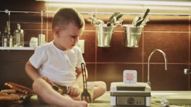 Genç çocuk ev mutfağında waffle makinesinde waffle yapıyor. Küçük çocuk masada oturuyor ve waffle makinesine zamanlayıcı takıyor.
