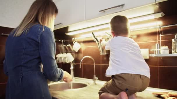 Petit garçon fils assis sur la table et aide maman dans l'évier de cuisine — Video