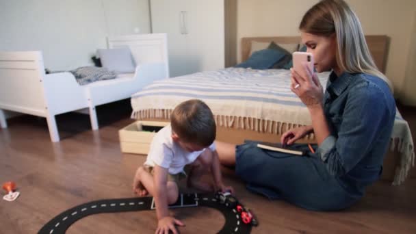 Busy woman talking on phone and making notes while her son plays cars on floor — Stock Video