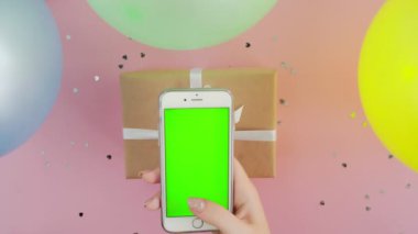 Kadının üst görüntüsü yeşil ekranlı, hediye kutusu pembe arka planda telefon kullanıyor.