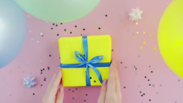 Mulher toca caixa de presente de aniversário de Natal envolto em papel amarelo com laço azul na mesa rosa — Vídeo de Stock