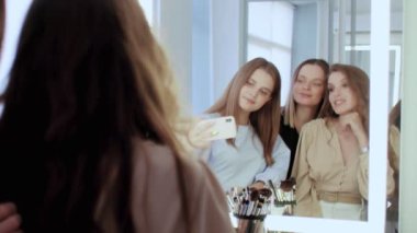Kendine güvenen beyaz makyajcı ve iki genç model güzellik stüdyosunda. Aynanın önünde selfie çekiyorlar.
