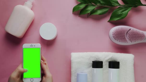 Kvinde banker og glider på hendes telefon med grøn skærm. Spa og kropspleje sammensætning. Wellness og skønhed behandling. Flat lay sortiment af lyserød baggrund. – Stock-video