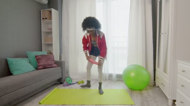 Ung afrikansk amerikansk kvinna hippy i färgglada sportkläder med lockigt afro hår gör stretching övningar med elastiskt band på benen — Stockvideo